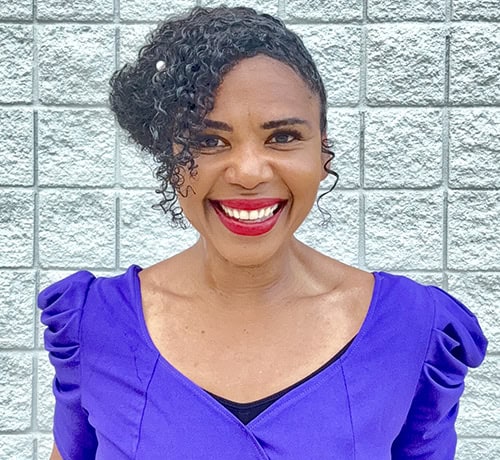 Smiling woman in purple top outside against a brick wall.